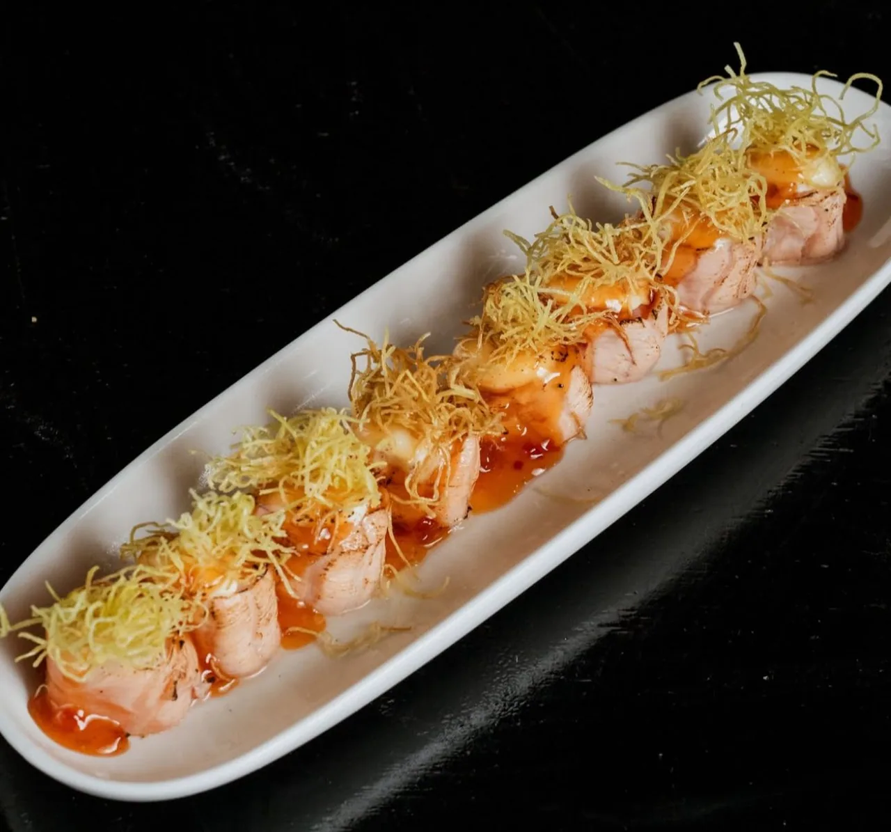 Hot roll de salmão maçaricado com fios crocantes, sobre travessa branca em fundo escuro